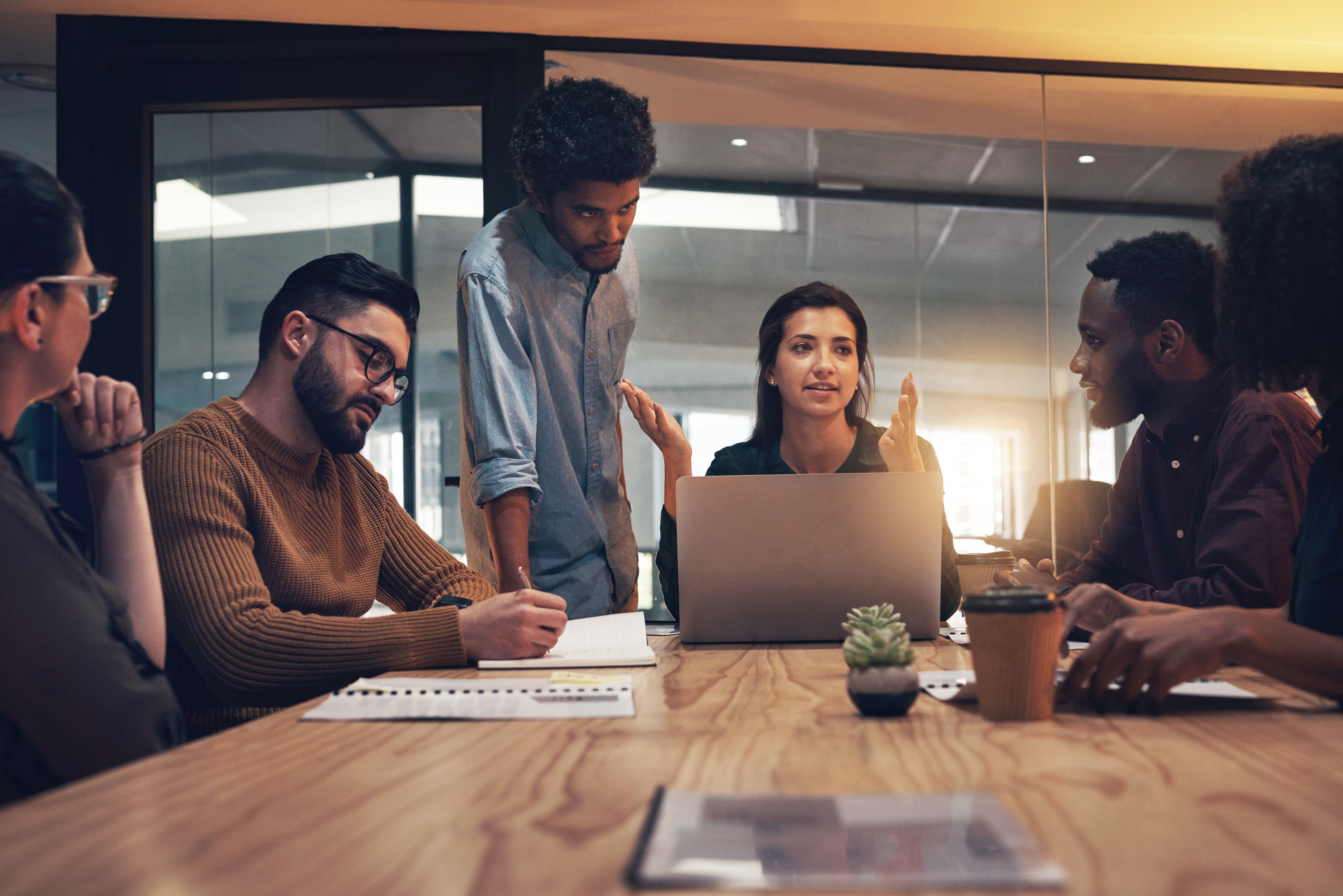 Three people in conversation, two seated at a wooden table, one sitting in front of a laptop and gesturing.