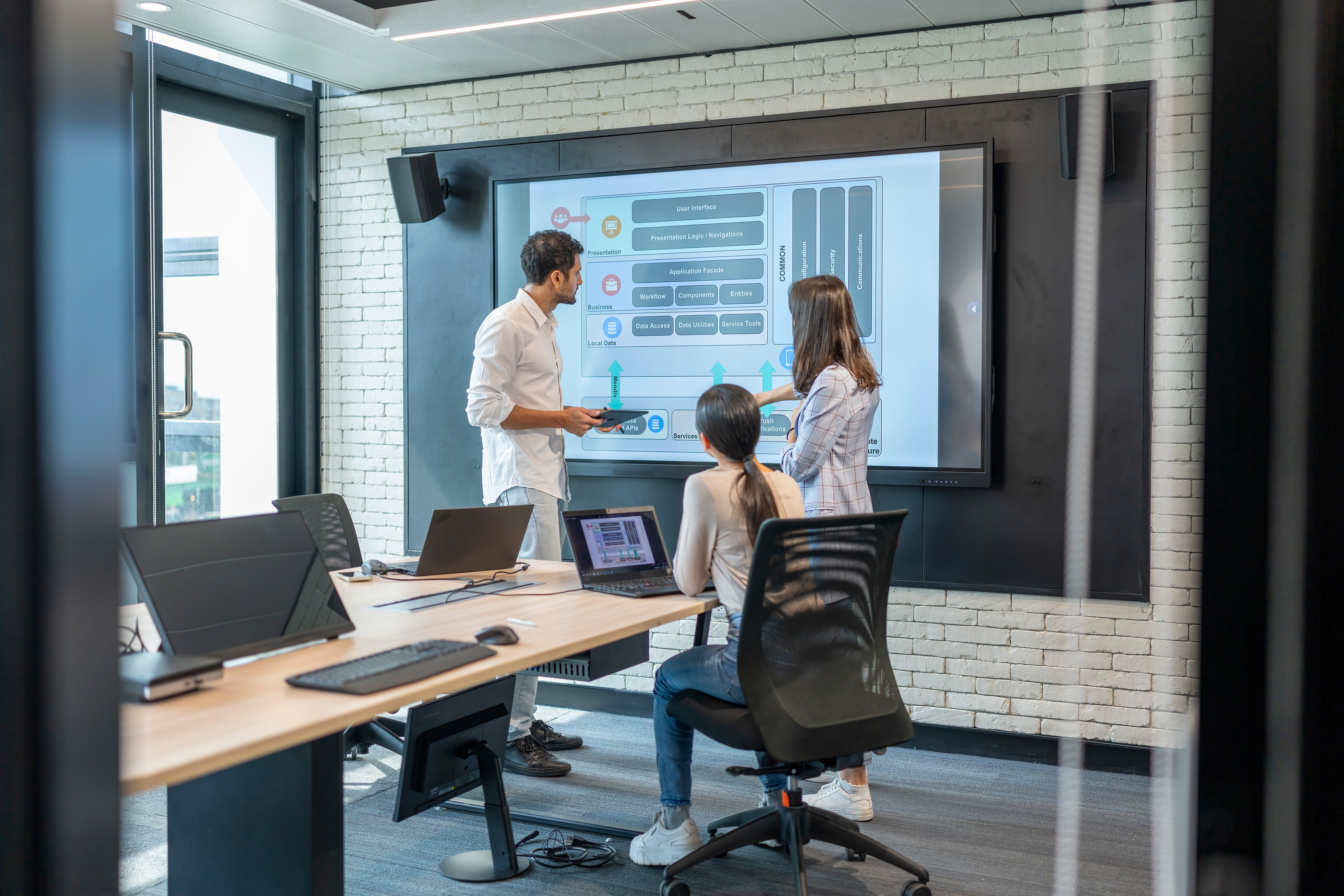 Drei Personen stehen in einem modernen Konferenzraum vor einem großen Bildschirm und besprechen ein technisches Diagramm. Zwei Laptops stehen auf dem Konferenztisch, während eine Person sitzt und zwei stehen. Die Atmosphäre wirkt konzentriert und kollaborativ.