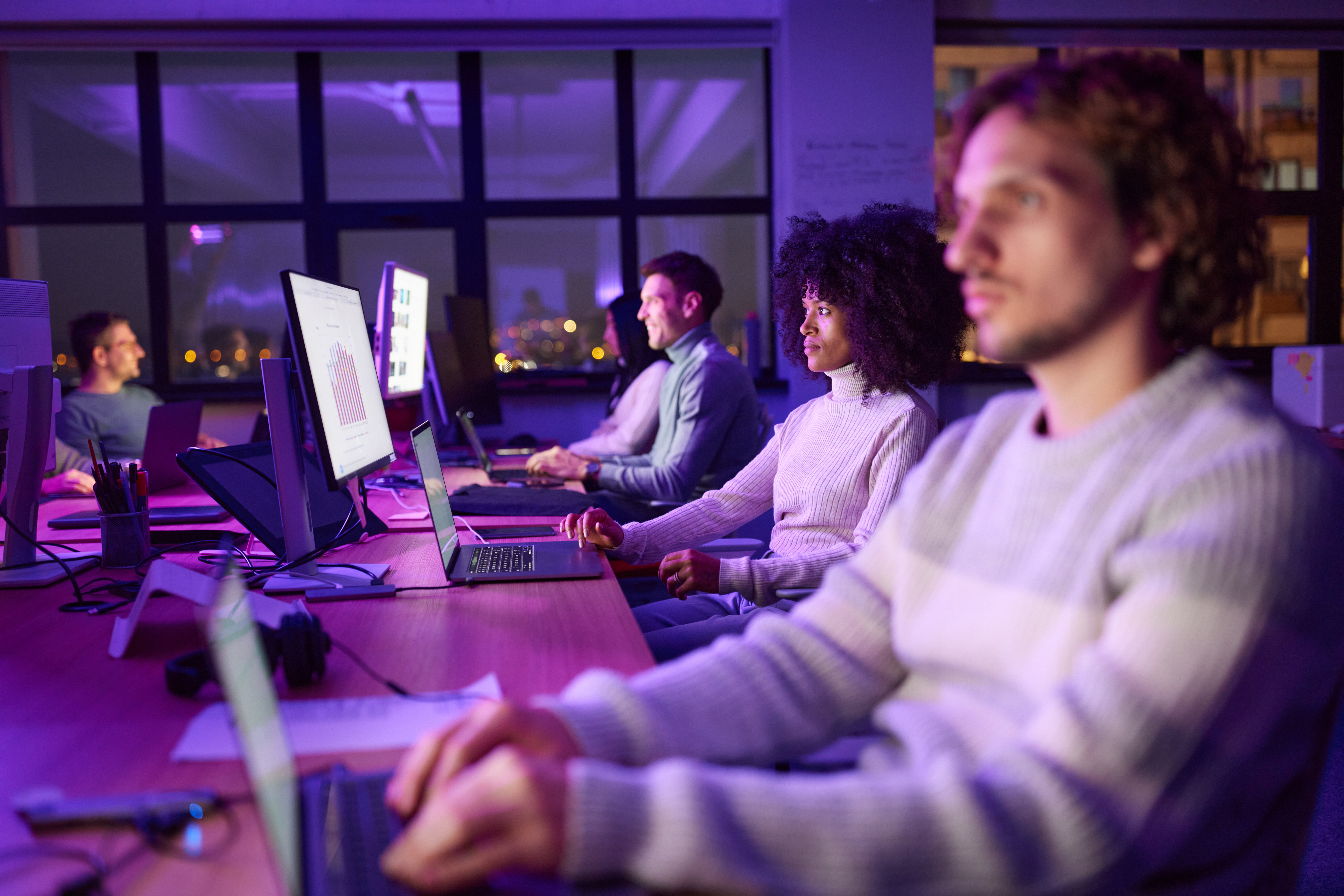 Colleagues work at computer monitors and laptops in a purple-lit modern office at night