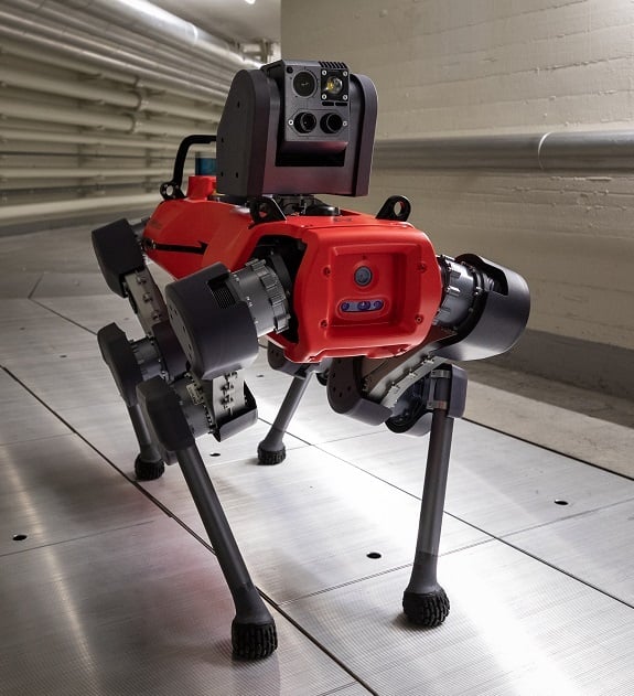 Eine hundeähnlicher Roboter mit rotem Korpus und schwarzen Beinen mit oben montierten Sensoren steht in einem Flur mit Rohren.