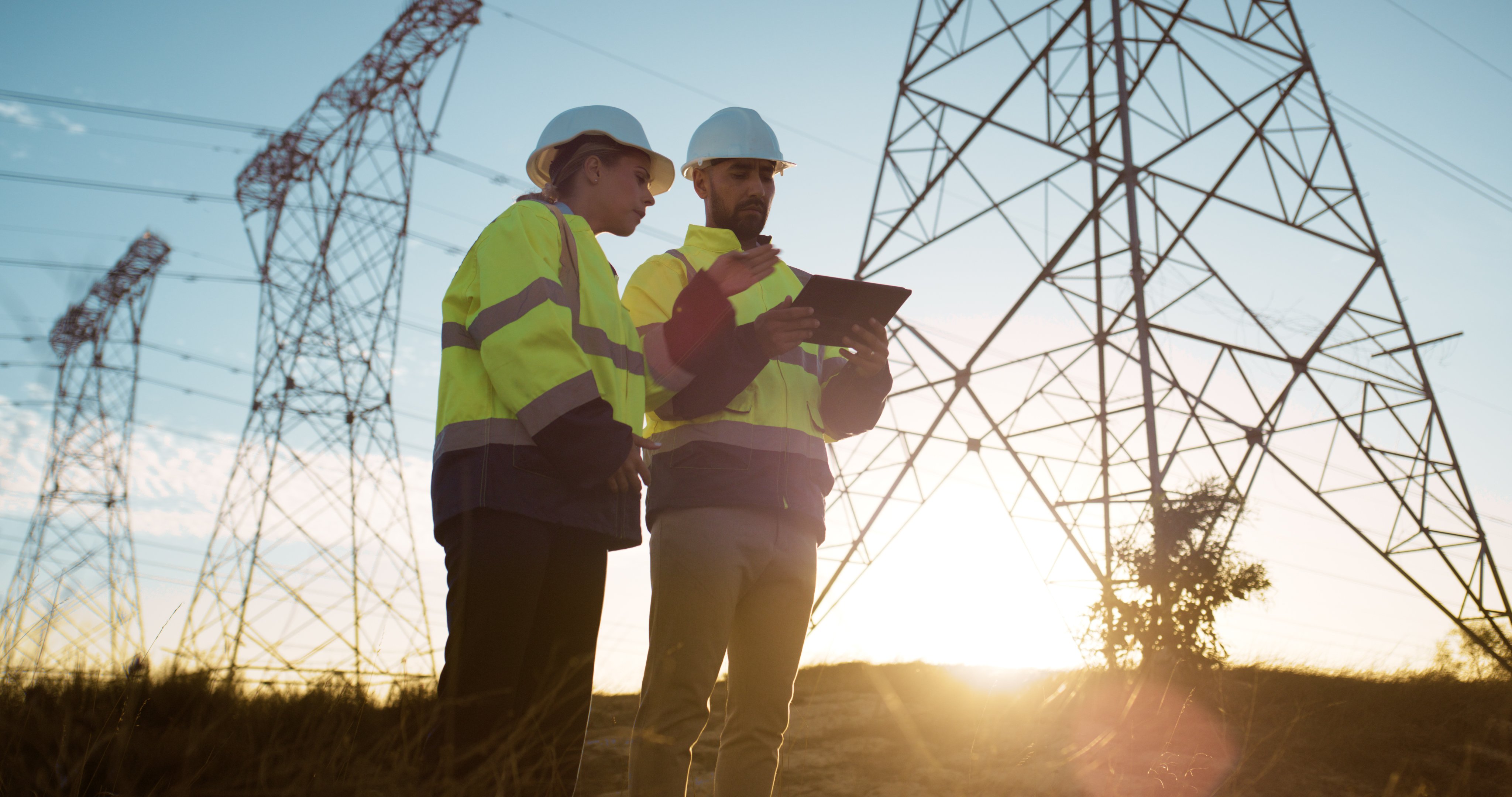 Two people in work clothes, holding an iPad, talk about asset management in the energy sector while the sun sets in the background.
